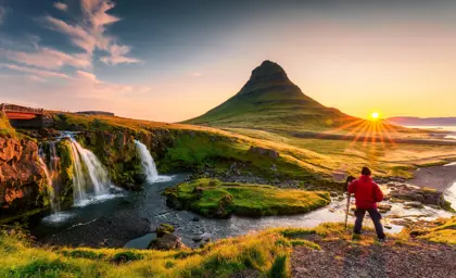 Hiker on tour around Iceland with trekking poles photographing the sunrise over Kirkjufell mountain and Kirkjufellsfoss waterfall in west Iceland.