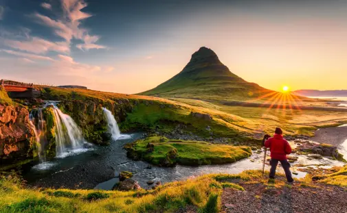 Hiker on tour around Iceland with trekking poles photographing the sunrise over Kirkjufell mountain and Kirkjufellsfoss waterfall in west Iceland.