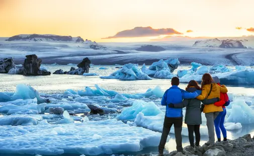 A group of travelers on a private glacier lagoon tour admiring the breathtaking Jökulsárlón Glacier Lagoon at sunset, with massive icebergs floating in the serene water under a golden sky in Iceland.