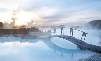 Misty sunrise at Blue Lagoon Iceland with steam rising over the geothermal spa and a wooden bridge reflecting in the tranquil blue waters.