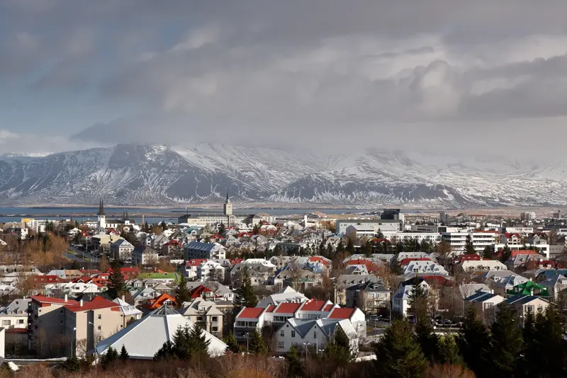 Panoramic view of Reykjavik under a snowy sky, featuring colorful houses, modern buildings, and the Esja mountain in the background.