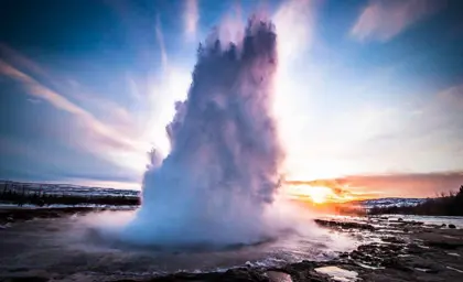 Strokkur Geyser erupting at sunrise in Iceland, showcasing a dramatic water plume against a colorful sky with a snowy winter landscape in the background.
