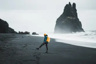 Young woman with yellow backpack and hat walks across Reynisfjara black sand beach toward dramatic rock formations close to Vík í Mýrdal.