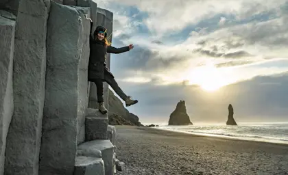 Female traveler posing playfully on basalt columns at Reynisfjara black sand beach at sunset in Iceland
