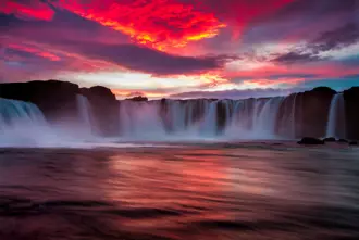 Dramatic view of Goðafoss Waterfall at sunset, with vivid red and purple sky reflecting on the flowing water, part of Iceland's natural wonders.