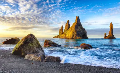 Golden sunlight illuminates the Reynisdrangar sea stacks at sunrise, with waves gently washing onto the black sand beach near Vík, Iceland.