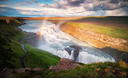 A rainbow arches over the powerful Gullfoss waterfall on a sunny day in Iceland's Golden Circle.