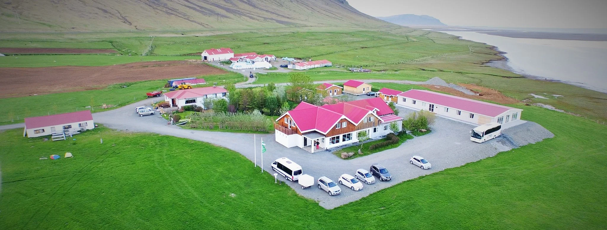Aerial view of Gerði Guesthouse, a cozy farmstay with red rooftops, nestled in green fields near the coastline, featured on the Blue Ice Cave and Glacier Lagoon tour.