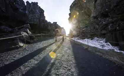 Visitors walking through a rocky pathway at Thingvellir National Park, Iceland, with sunlight breaking through the cliffs, showcasing the park's dramatic landscapes and historical significance.