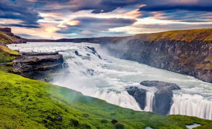 Scenic view of Gullfoss Waterfall during a vibrant sunset, with tourists walking along the green path as part of the Golden Circle tour in Iceland.