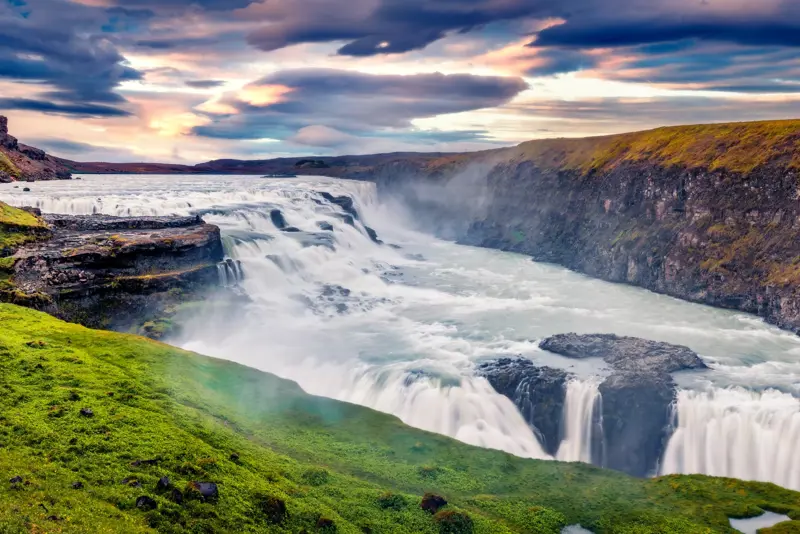 Scenic view of Gullfoss Waterfall during a vibrant sunset, with tourists walking along the green path as part of the Golden Circle tour in Iceland.