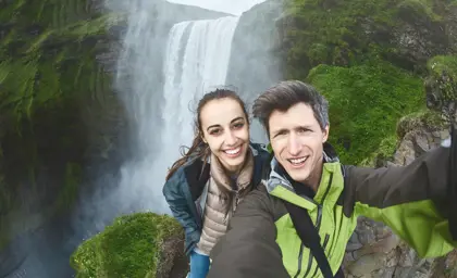 Happy couple taking a selfie in front of Skogafoss waterfall during their Iceland hiking adventure