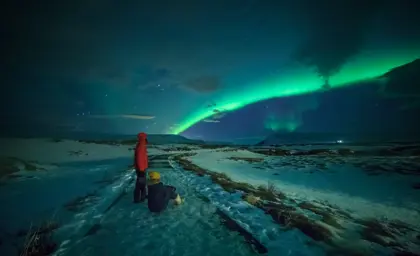 Couple watches the northern lights on a 3 day northern lights tour in Iceland under a vivid green aurora arc in the snowy Icelandic countryside at night.