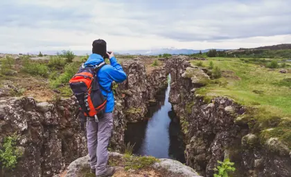 Male hiker in a blue jacket and backpack observing a dramatic rift valley filled with clear water at Thingvellir National Park in Iceland, surrounded by moss-covered rocky cliffs and green fields under a cloudy sky.