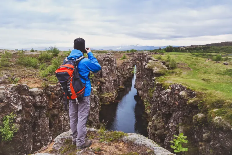 Male hiker in a blue jacket and backpack observing a dramatic rift valley filled with clear water at Thingvellir National Park in Iceland, surrounded by moss-covered rocky cliffs and green fields under a cloudy sky.