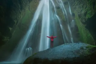 Woman with arms raised under Gljúfrabúi waterfall, surrounded by mist and mossy cliff walls in Iceland.