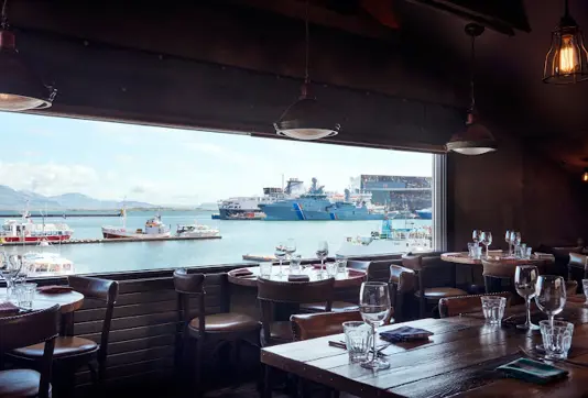 View of Reykjavik harbor from inside Kopar Restaurant, featuring elegantly set dining tables and a backdrop of boats and industrial ships under a clear sky.