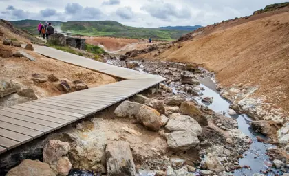 A scenic wooden boardwalk meandering through the colorful geothermal area of Seltún in Iceland, with steam vents and rugged terrain visible under an overcast sky.