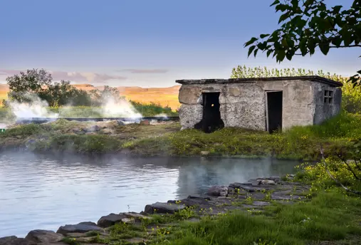 Secret Lagoon original stone bathhouse in gamla laugin in Flúðir, Iceland, surrounded by natural geothermal steam and wildflowers on a serene summer evening.