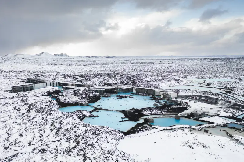 Aerial view of the Blue Lagoon in Iceland during winter, surrounded by snowy lava fields, with geothermal pools and spa facilities nestled in a serene, icy landscape.