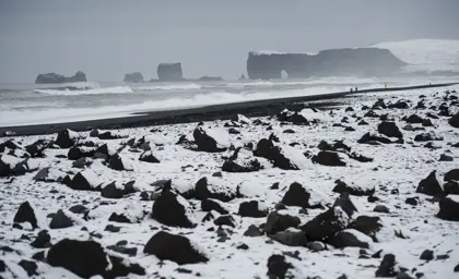 Snow-covered Reynisfjara Black Beach with rugged volcanic rocks and distant view of Dyrhólaey in Iceland during winter.
