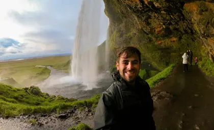 Happy traveler taking a selfie behind Seljalandsfoss waterfall on the mossy trail in Iceland.