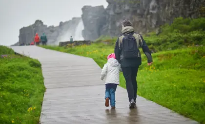 Father and young daughter walking hand in hand along a wooden boardwalk through Thingvellir National Park in Iceland, surrounded by lush green grass and dramatic cliffs.