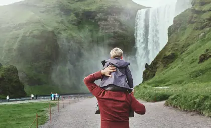 Father carrying son on shoulders while walking towards the misty Skógafoss waterfall in southern Iceland on a family trip.
