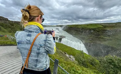 Tourist woman capturing the powerful Gullfoss waterfall in Iceland on a cloudy summer day from the viewing platform.