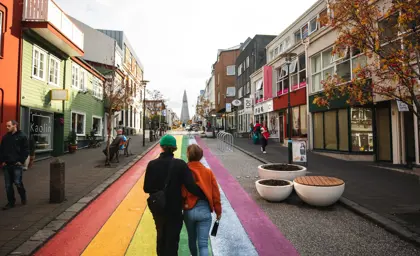A couple walking along the colorful rainbow-painted street leading to Hallgrímskirkja in Reykjavík, Iceland, surrounded by vibrant buildings and autumn trees. Part of a Reykjavík city tour experience.