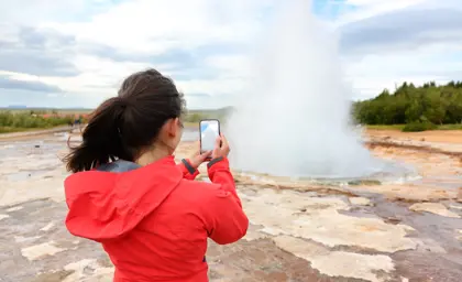 Female traveler in red jacket photographing Strokkur geyser eruption in Iceland's Golden Circle geothermal area.