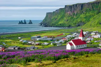 Scenic view of Vík í Mýrdal, showcasing colorful houses, a church with a red roof, and Reynisdrangar sea stacks in the background, highlighting Iceland's natural beauty.