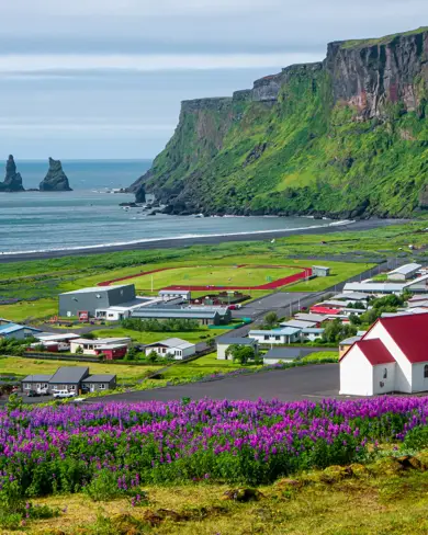 Scenic view of Vík í Mýrdal, showcasing colorful houses, a church with a red roof, and Reynisdrangar sea stacks in the background, highlighting Iceland's natural beauty.