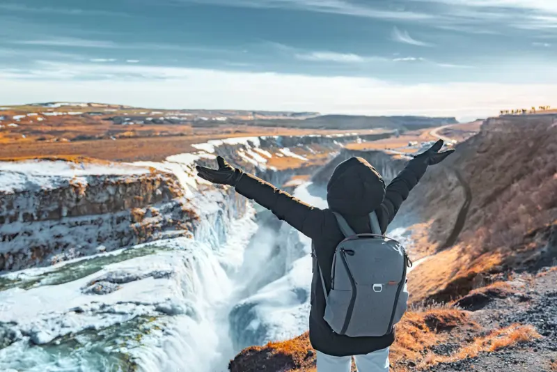 Joyful female tourist raising her arms while admiring the powerful Gullfoss waterfall surrounded by snow in Iceland.