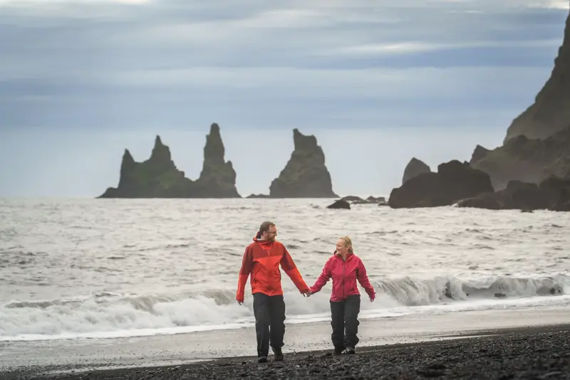 Smiling couple walks hand-in-hand on Reynisfjara black sand beach with Reynisdrangar basalt sea stacks in the background.