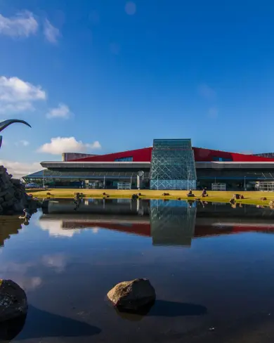 View of Keflavík International Airport in Iceland, featuring its modern architecture and iconic reflective sculpture by a calm pond on a sunny day.