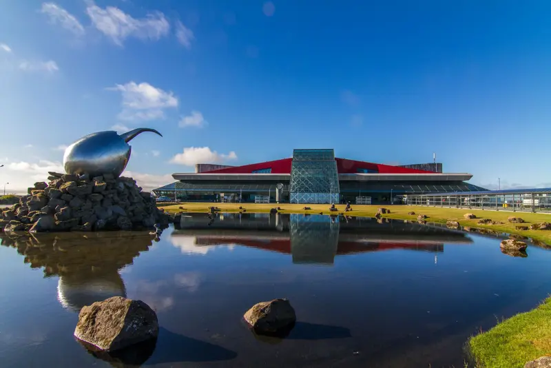 View of Keflavík International Airport in Iceland, featuring its modern architecture and iconic reflective sculpture by a calm pond on a sunny day.