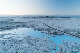 Winter aerial view of the Blue Lagoon geothermal spa, surrounded by volcanic lava fields and snow, showcasing the stunning icy blue water and a winding road leading to the spa in Iceland.