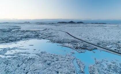 Winter aerial view of the Blue Lagoon geothermal spa, surrounded by volcanic lava fields and snow, showcasing the stunning icy blue water and a winding road leading to the spa in Iceland.