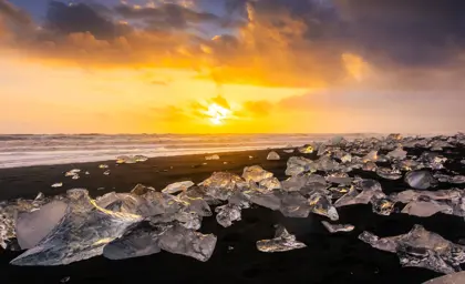 Glacial ice chunks scattered across the black sand of Diamond Beach in Iceland glow under a vibrant orange sunset sky, with waves crashing in the background.