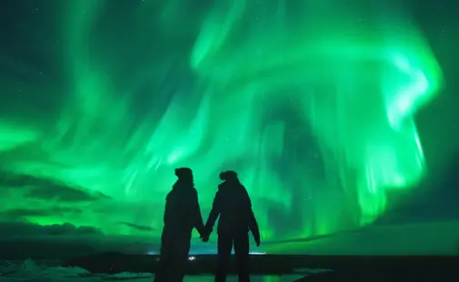 Couple holding hands under dazzling green northern lights in Iceland, reflecting in a still icy lagoon for a magical winter experience.