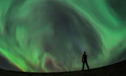 Lone traveler with headlamp on a 5 day northern lights tour in Iceland gazing at star-filled sky and swirling northern lights.