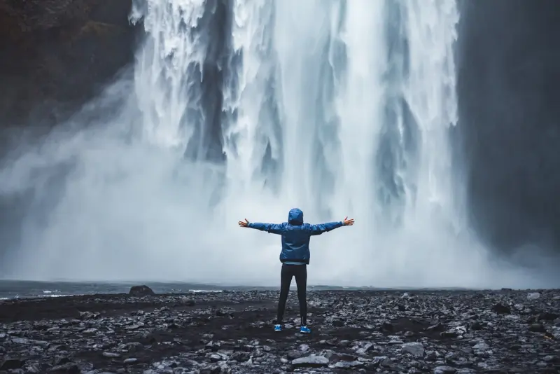 A person in a blue jacket stands with arms outstretched in awe before the powerful Skogafoss waterfall in Iceland, surrounded by mist and rocky terrain.
