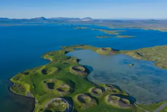 Aerial view of Skútustaðir pseudo craters on the shores of Lake Mývatn in North Iceland, showcasing unique volcanic landscape formations surrounded by lush green grass and pristine blue waters under a clear sky.