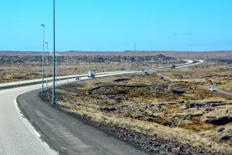 A view of Reykjanesbraut highway cutting through the rugged lava fields of Reykjanes Peninsula under a bright blue sky, showcasing the unique volcanic terrain of Iceland.