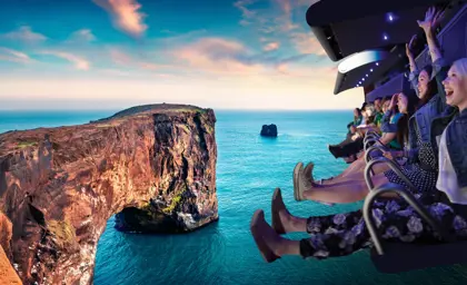 Visitors flying above Dyrhólaey cliff on the FlyOver Iceland experience, with a breathtaking view of the Icelandic coastline and rock formations against a clear sky.