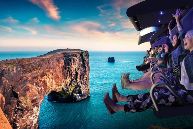 Visitors flying above Dyrhólaey cliff on the FlyOver Iceland experience, with a breathtaking view of the Icelandic coastline and rock formations against a clear sky.