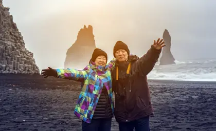 Senior couple smiling and stretching their arms at Reynisfjara black sand beach in Vík, Iceland, with dramatic sea stacks and basalt cliffs in the background.