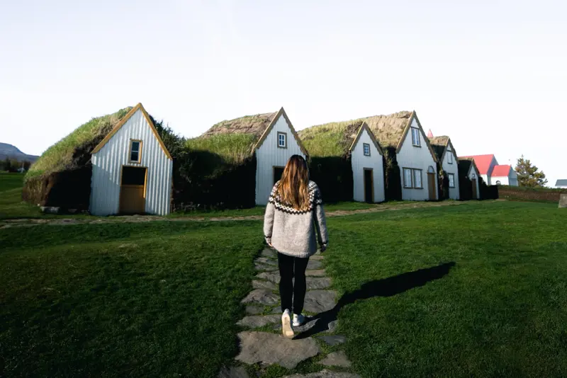 Glaumbær turf house farm and museum behind a woman walking towards it surrounded by green grass and clear skies. 
