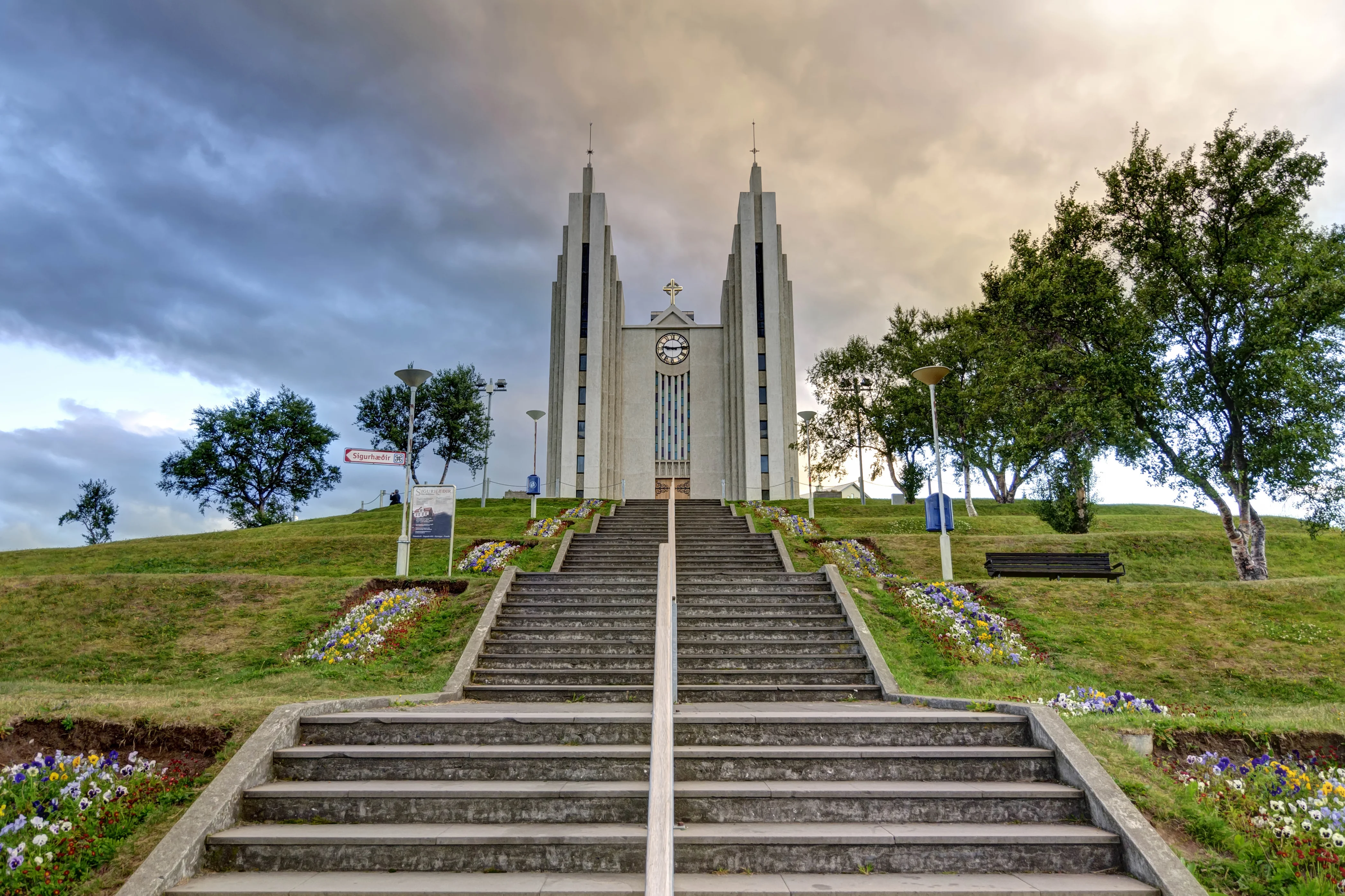 Akureyri Church (Akureyrarkirkja) on a hill with its iconic concrete structure and steps leading up, surrounded by trees and gardens.
